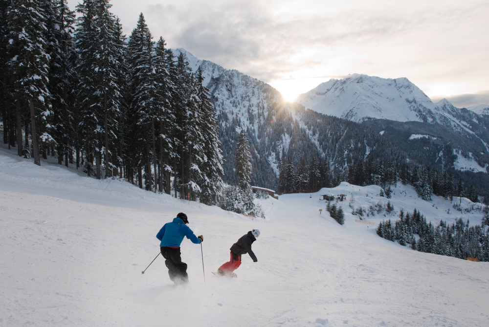 zwei Personen auf Ski und Snowboard auf der Piste mit Bäumen am Rand und Bergen im Hintergrund